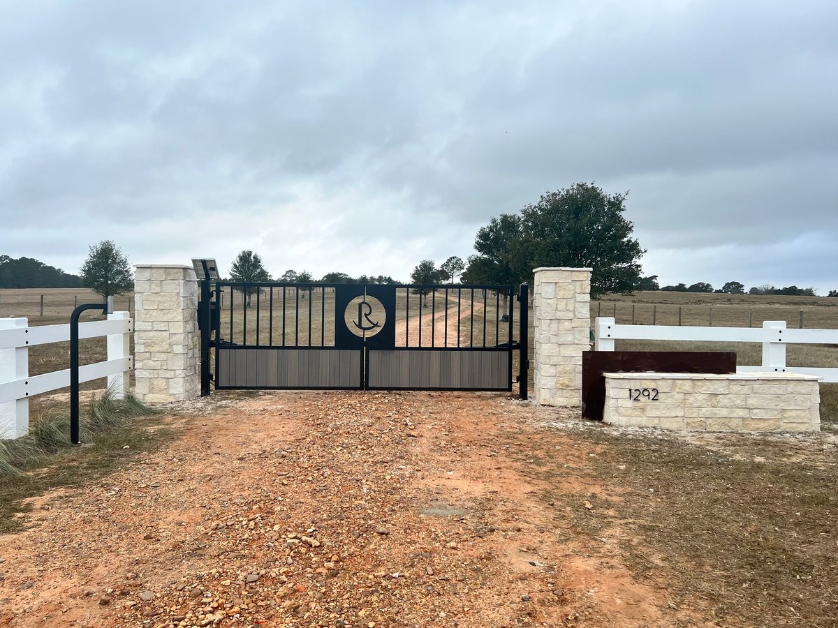 Custom black metal driveway gate with stone pillars and a monogram on a rural property