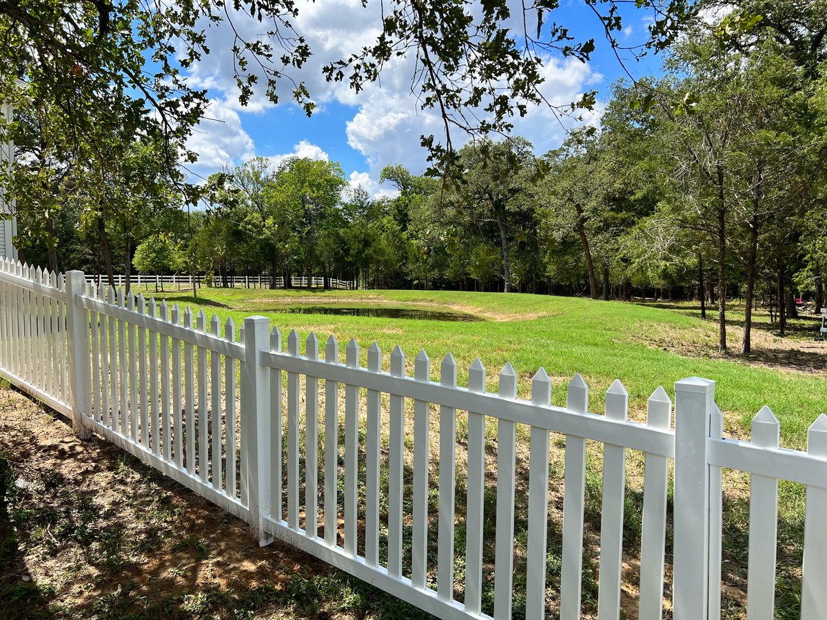 White picket fence lining a green pasture with trees under blue sky in Brookshire TX