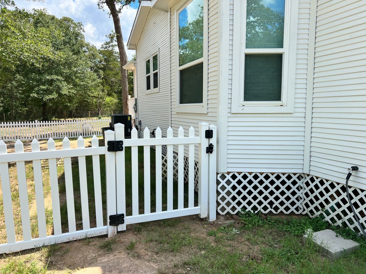 White picket fence with black gate in front of a light-colored home Brookshire TX