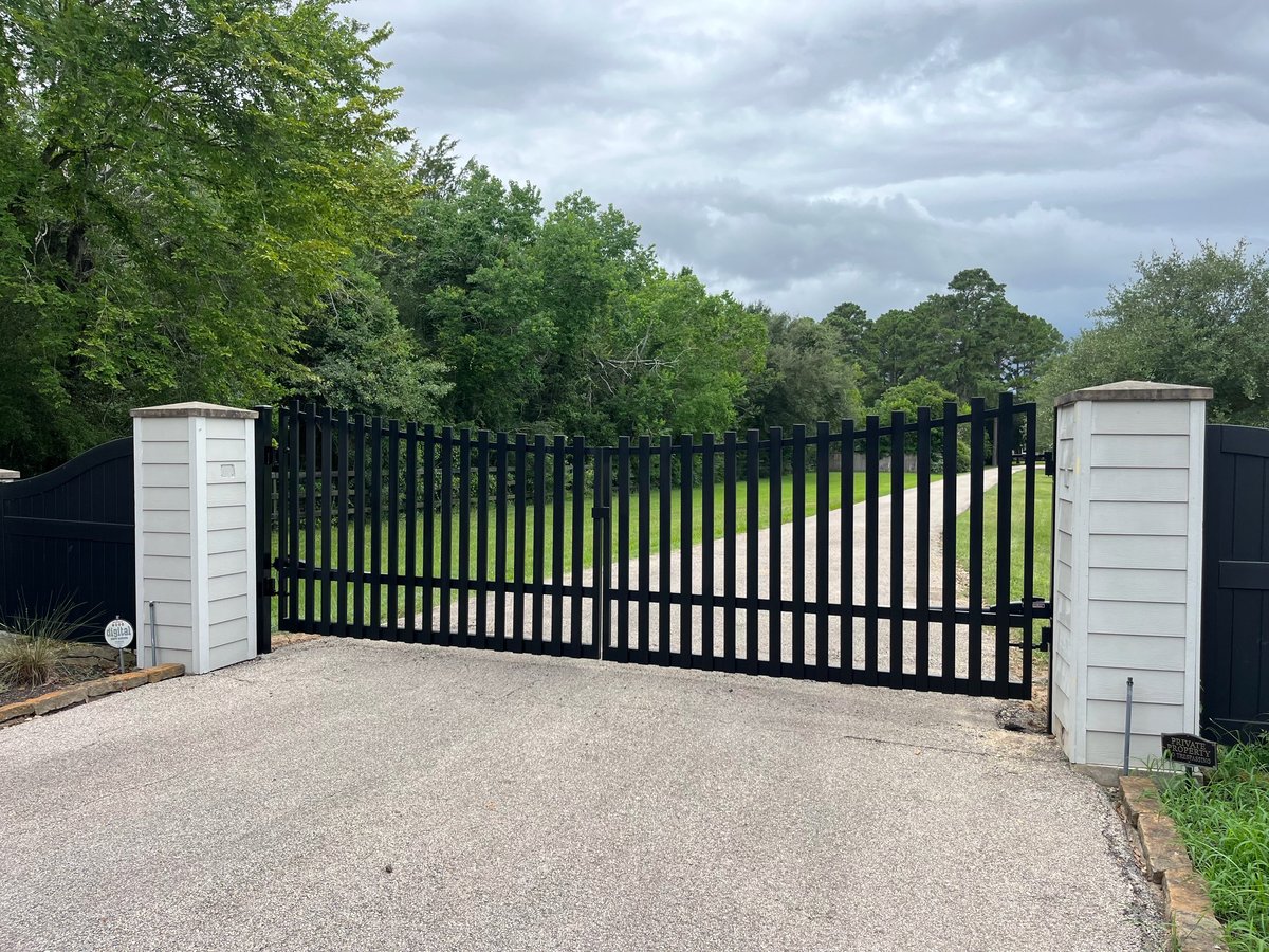 Black metal driveway gate with white pillars on a gravel path surrounded by green trees
