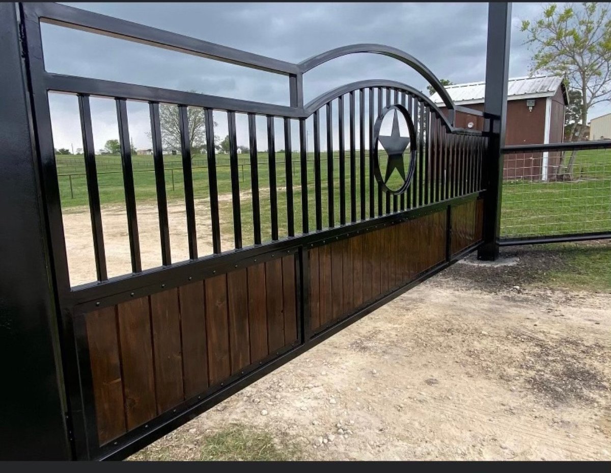 Black metal farm gate with wooden lower panels and curved top design with a Texas star, partially open in a rural setting with green fields